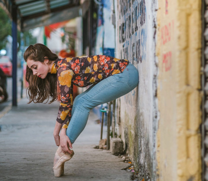 Woman wearing jeans and floral top and ballet pointe shoes leaning against wall in Rio de Janeiro