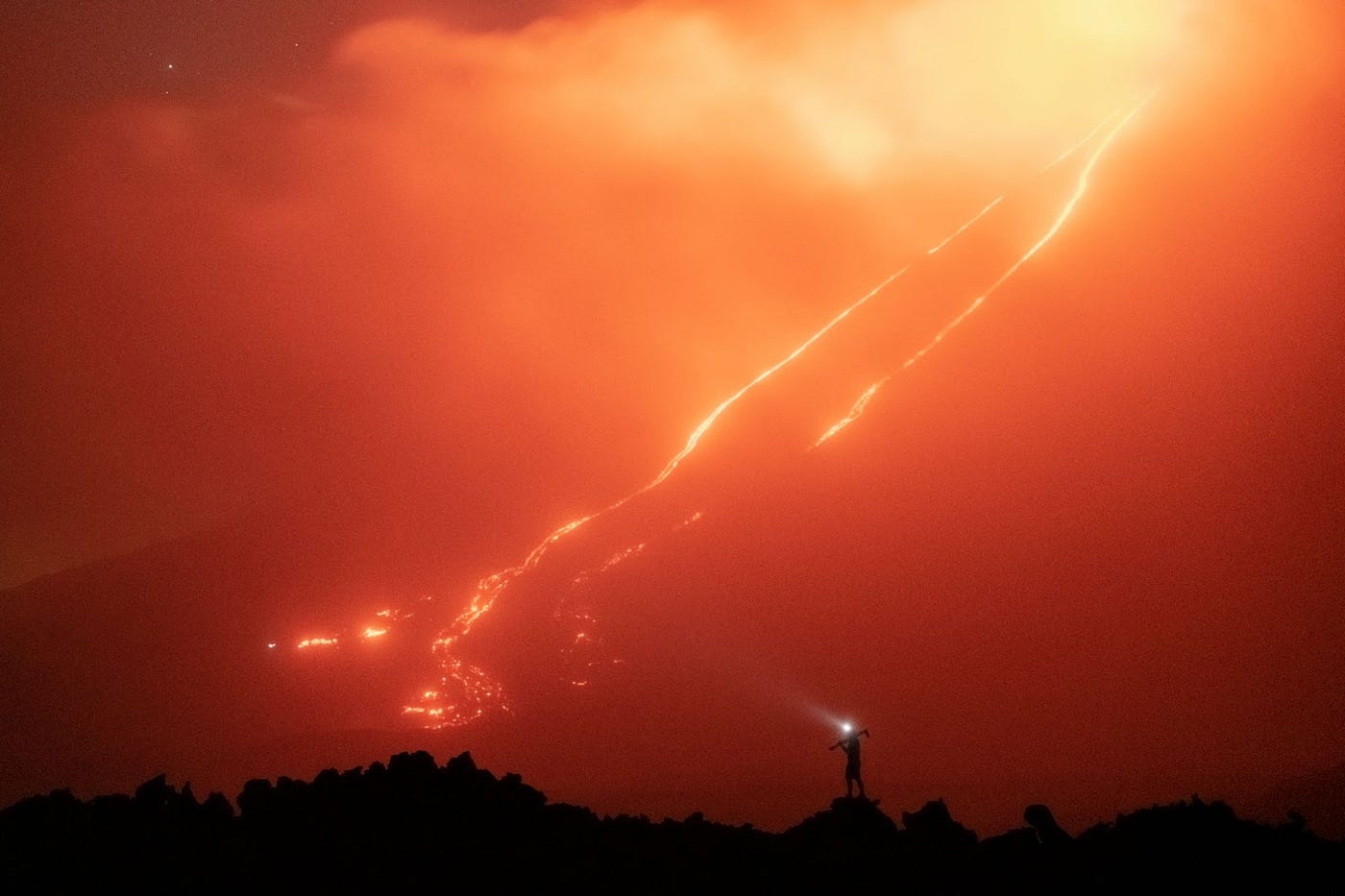 Silhouette of a photographer with a headlamp and tripod standing in front of an active volcano with lava flow.